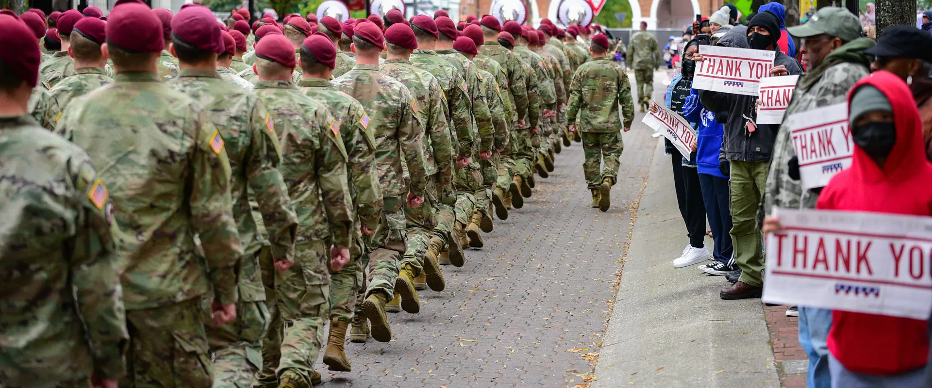 Soldiers in maroon berets march down a street lined with people holding Thank You signs